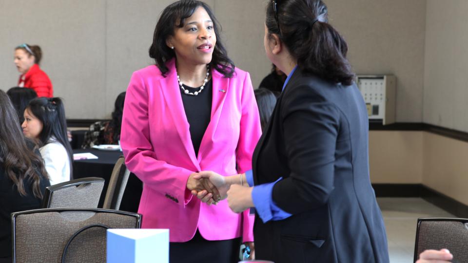 Two women shaking hands, one wearing a bright pink blazer, the other wearing a black blazer and facing away from the camera