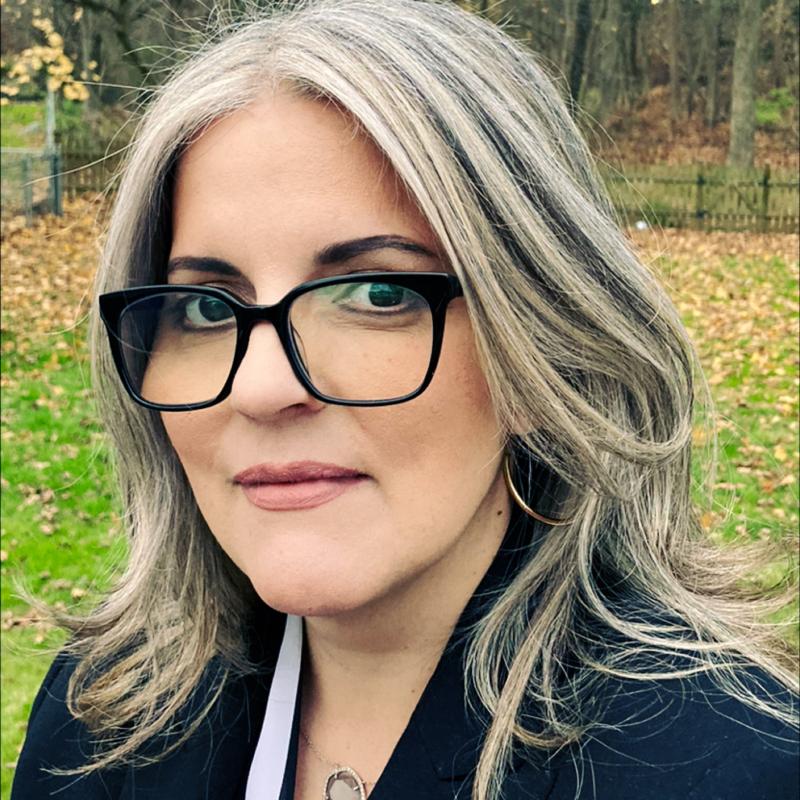 Head shot of Saily Avelenda wearing a black jacket ad black glasses standing against a background of trees
