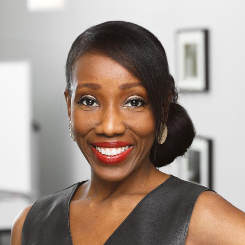 Head shot of Tara Dowdell wearing a black v-neck shirt against a wall with framed photos