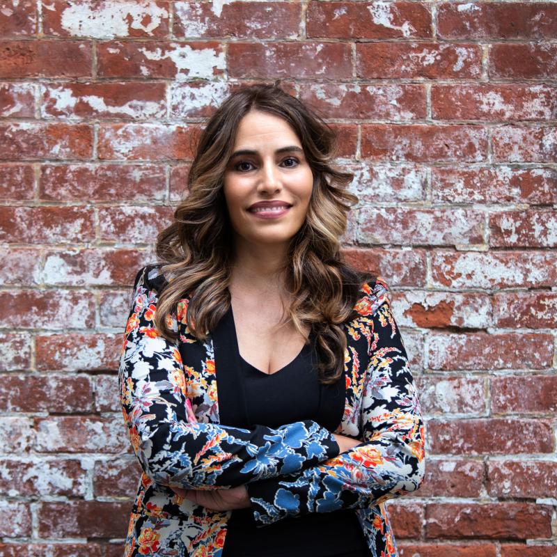 Head shot Senator Teresa Ruiz wearing a multi-color blazer and black shirt, standing with arms crosses against a brick background