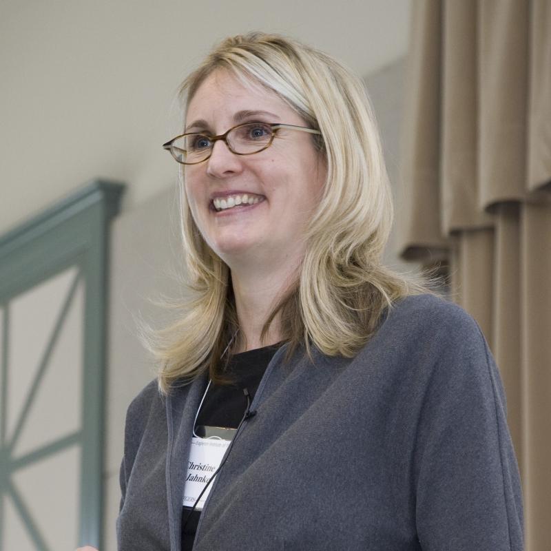 Woman with blond hair and glasses wearing a black shirt and gray jacket.