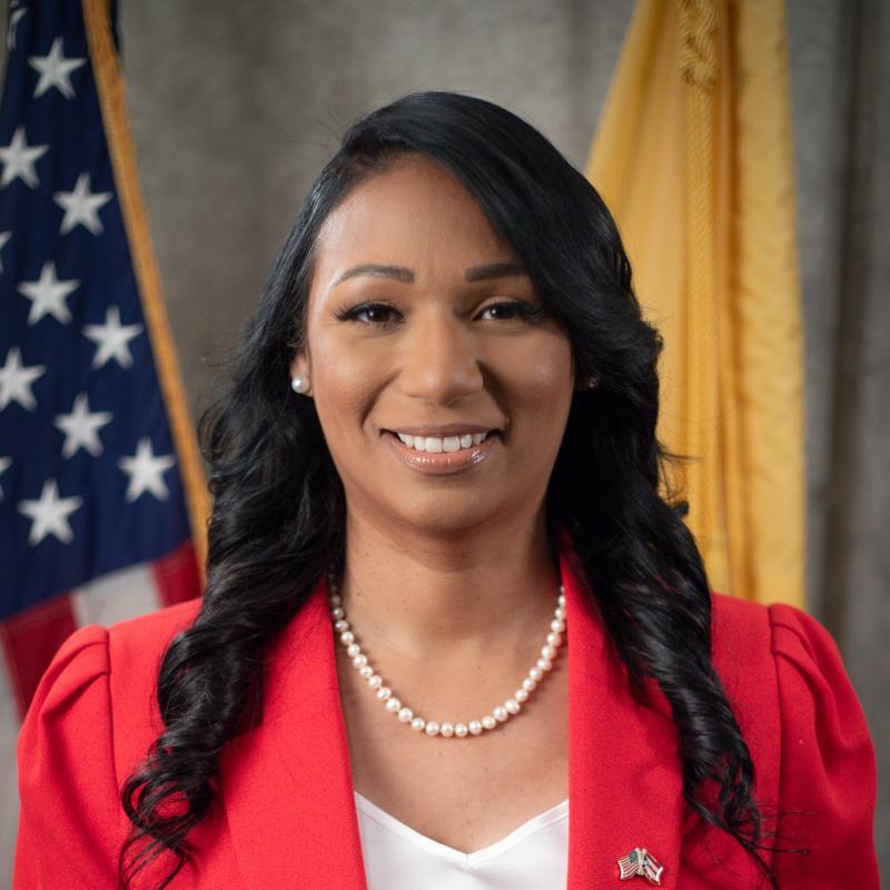 A photo of a brown skinned woman with black hair. She is wearing a pink blazer and sitting in front of two flags
