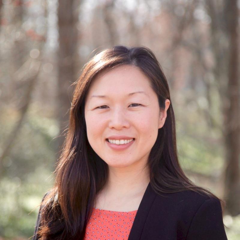 A woman in an orange shirt is standing outside, near the woods, smiling at the camera