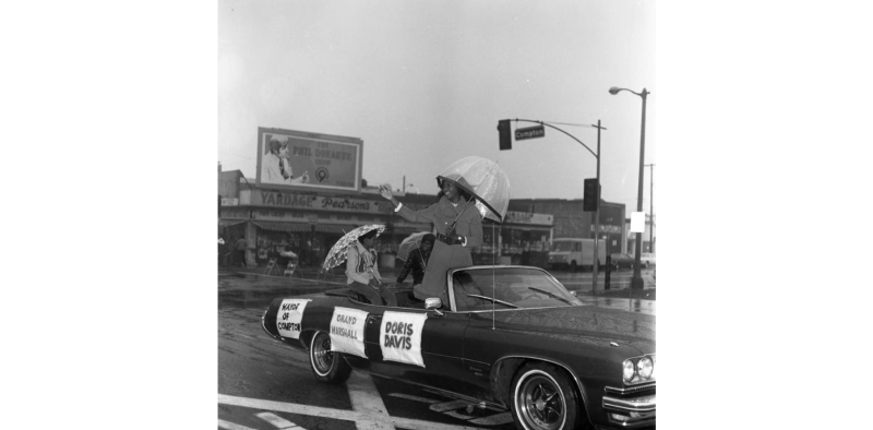 Doris Davis riding in convertible in rain as parade Grand Marshall.