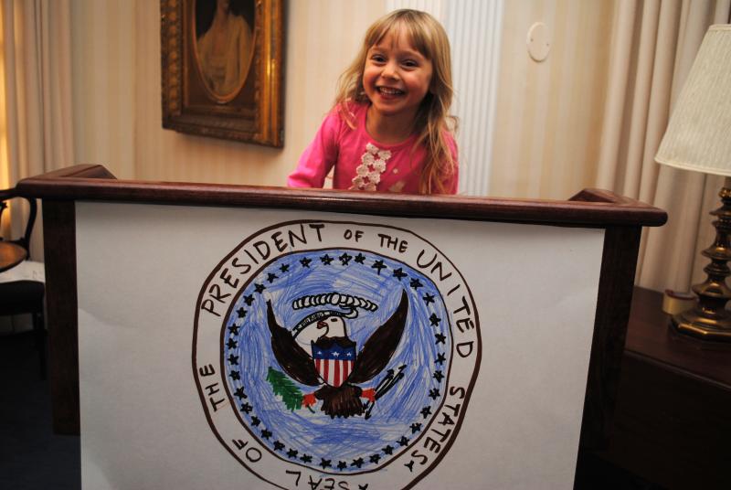 Girl with blond hair and pink shirt standing behind wooden podium with a hand-drawn President of the United States seal