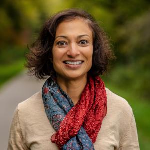 Paru Shah in a beige shirt with a blue and red scarf in front of a blurred outdoor background