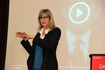 Chris Jahnke in front of TV screen, wearing black blazer, blue sweater, white collar shirt, glasses