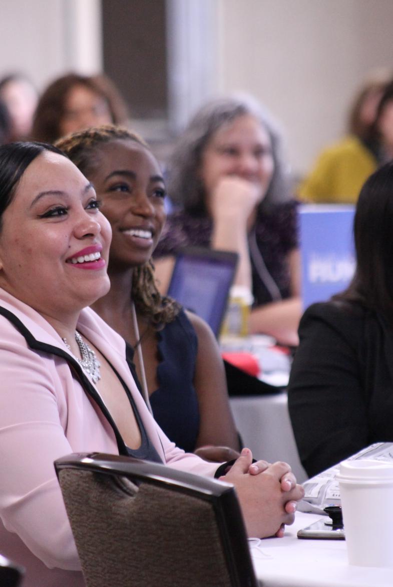 Women of different ages and races smile as they sit at tables in a conference room