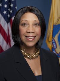 Sheila Y. Oliver smiles in black suit in front of American and New Jersey flags